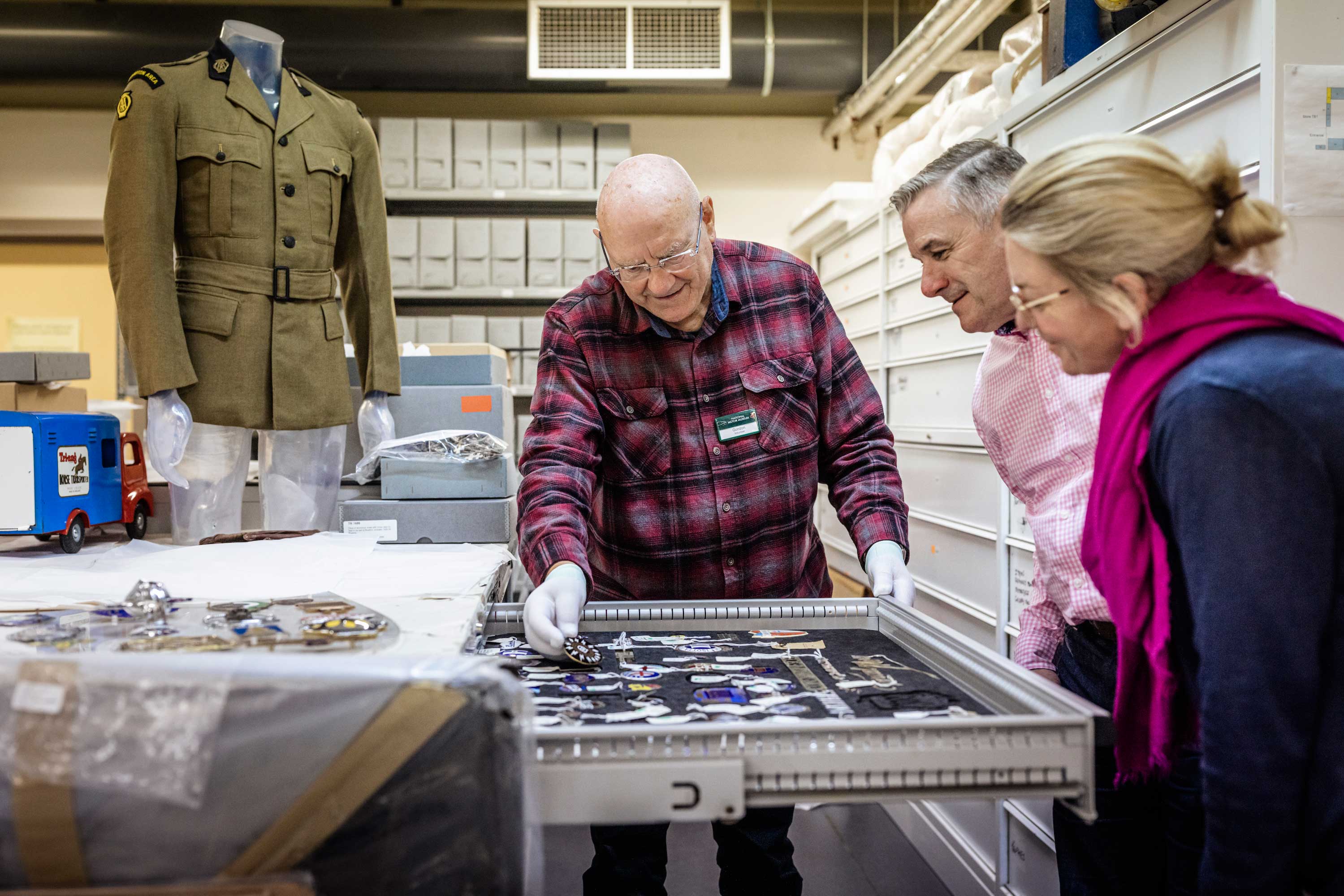 a man and woman being shown a drawer of car badges by a male member of staff in the object storeroom