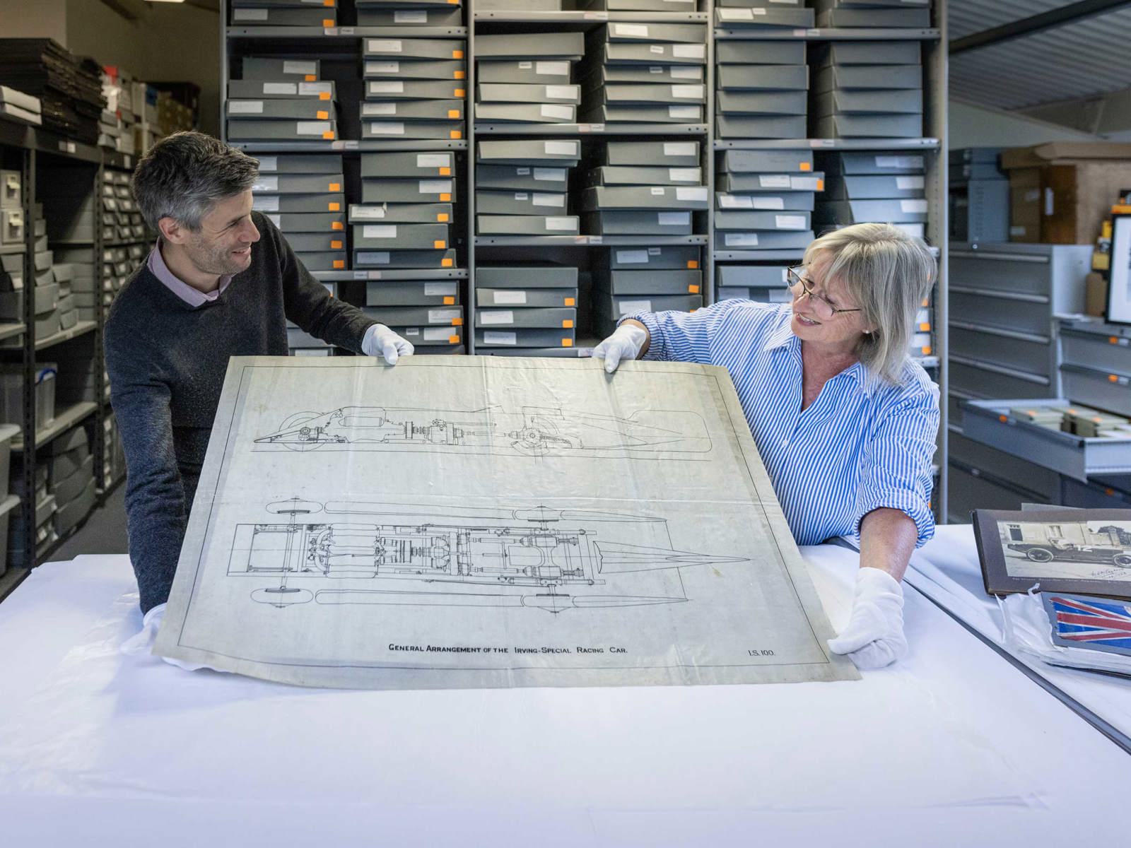 two members of curatorial staff hold up an engineering plan of Golden Arrow land speed vehicle