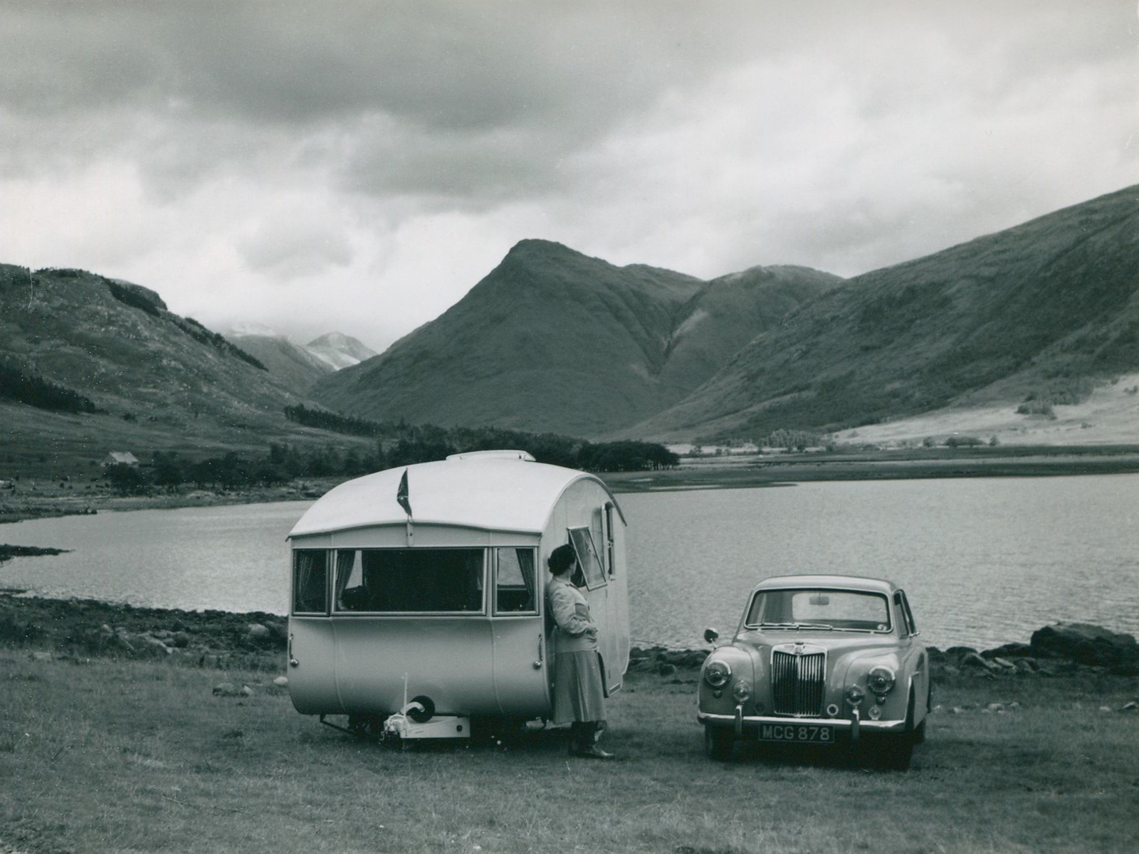 Hutchings Caravan and car parked near a lake, woman standing by the caravan